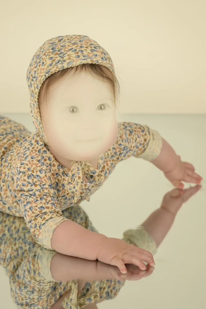 Baby wearing a floral bonnet and matching romper crawls on a reflective surface, showcasing the coordinated outfit's print.