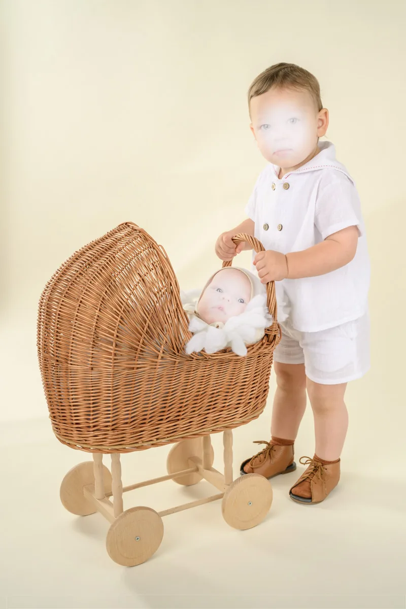A toddler in a white outfit stands beside a wicker wheeled bassinet, resting a hand on the handle as an infant looks out from inside.